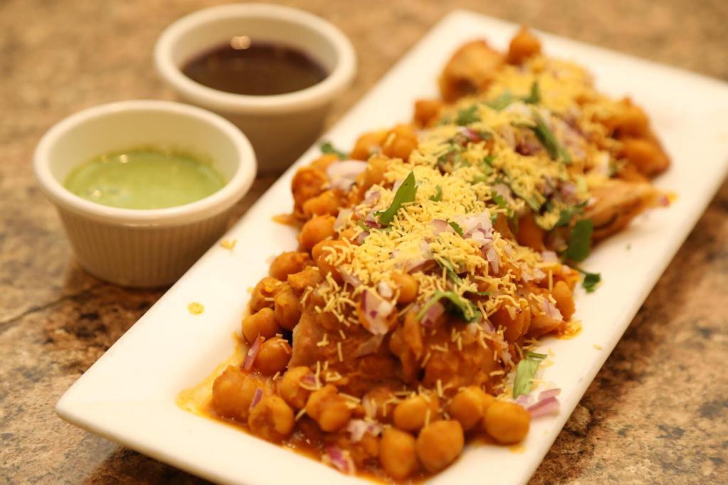 Samosa Chaat · Crispy samosa crumble in a spicy chickpeas base, topped with mint,tamarind and yogurt chutneys and garnished with onions and cilantro.