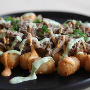 Loaded Tots · Crispy fried tots topped with kalua pig, spicy mayo, wasabi aji aioli, garnished with toasted sesame, green onions, + garlic chips.