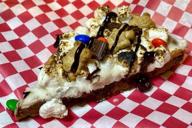 S'mores Cookie Slice · Chocolate chip cookie base, topped with vanilla buttercream, toasted with marshmallows, teddy grahams, M&M's, and drizzled with chocolate syrup.