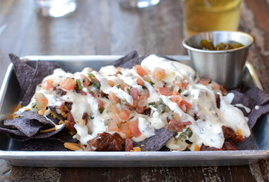 Brisket Chili Nachos · Gluten-free blue corn tortilla chips topped with smoked queso, brisket chili, sharp cheddar, house made pico de gallo and finished with cilantro lime sour cream served with candied jalapeños on the side.