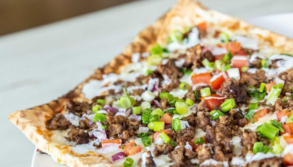 Kafta Beef Pizza · ground beef mixed with minced onions, jalapenos, parsley, Mediterranean seasoning, pomegranate molasses, tomatoes, fire-roasted jalapeno peppers, pine nuts, mozzarella, drizzled seasoned yogurt sauce and thin lavash crust
