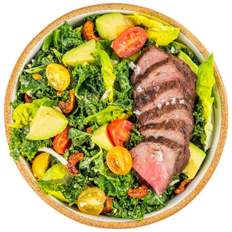 Steak Kale Caesar Salad · Cashew Kale Caesar, farm greens with mint, sliced peppercorn steak with grilled onion, avocado, shaved cauliflower, cherry tomatoes, spicy cashews, toasted breadcrumbs, and  more cashew Caesar dressing on the side. Contains wheat/gluten, tree nuts (cashew).