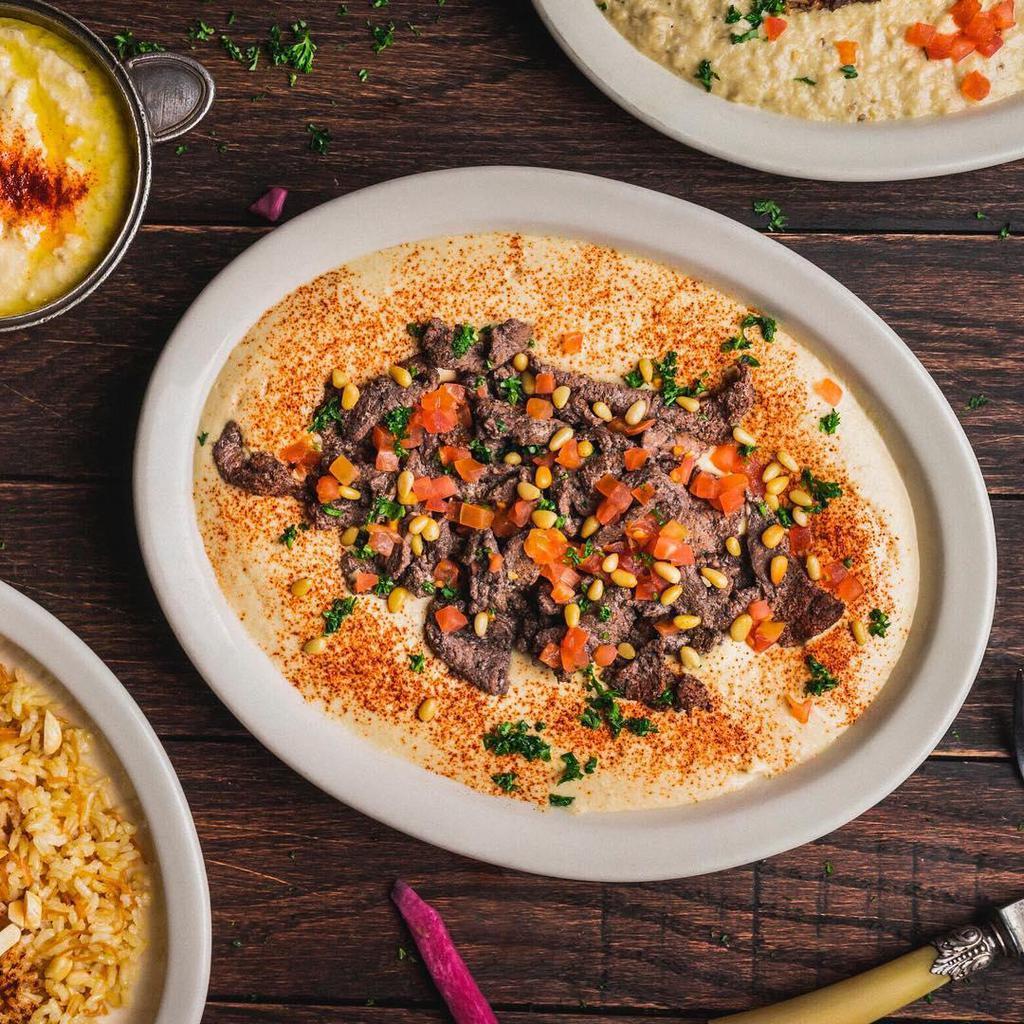 Hummus Shawarma Specialty Plate · Our award-winning hummus topped with grilled strips of seasoned beef sirloin, lightly fried pine nuts, diced tomatoes, and chopped parsley. Nuts and gluten-free.