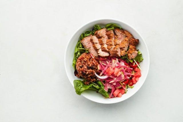 Carne Asada Bowl · Grass-fed sirloin steak with HB achiote marinade, pinto and black beans mixed with Himalayan ruby rice, mixed greens, watermelon, watermelon radishes, red onions, red chilies, cilantro. Gluten free.