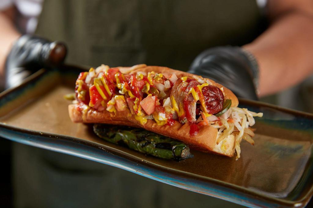 The MVP Sonora Dog · Applewood bacon wrapped hot dog on griddled bun with griddled onions, peppers, pickled cabbage slaw, tomato jalapeno relish, mayo, mustard, and tapatio ketchup.