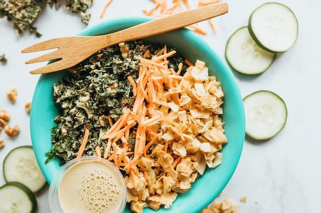 Marinated Kale Salad · Marinated kale tossed with carrots, cucumbers, and zesty serrano dressing. Topped with kale chips, coconut maple chips, and walnuts.
