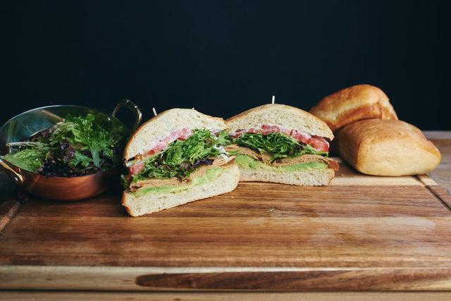 The West Coast Tofurky Sandwich · Oven-roasted tofurky, avocado, mixed greens, tomato, red onion, vegan basil aioli and lemon vinaigrette on a ciabatta. Vegan.