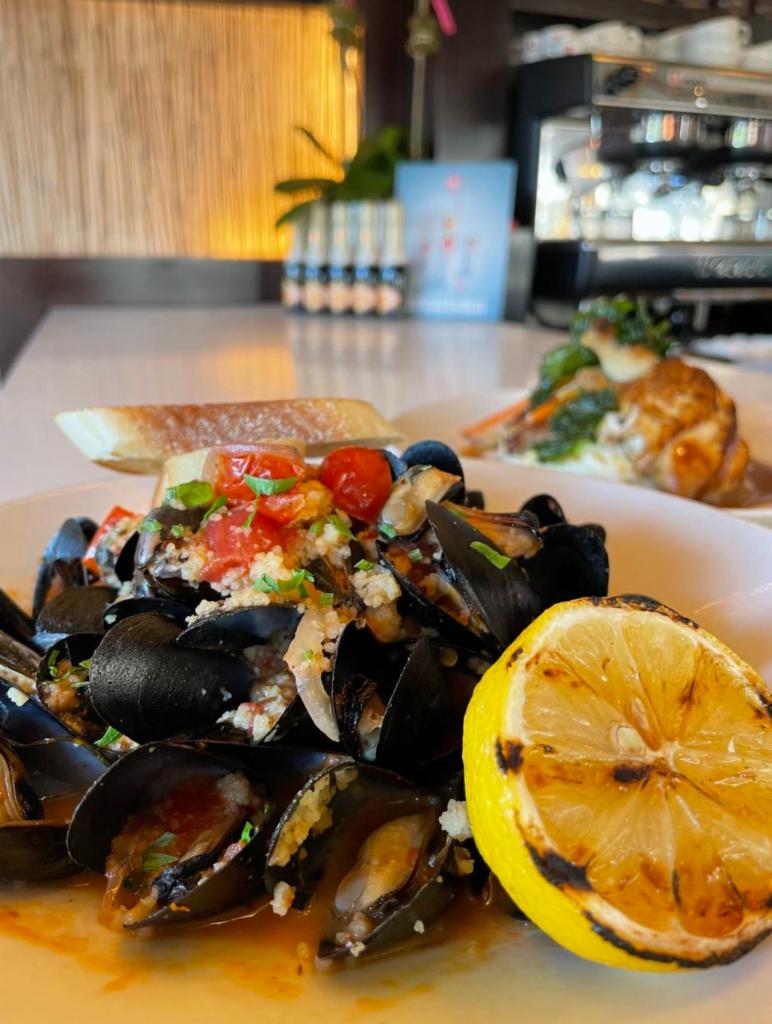 Mussels · steamed in a spicy and garlic tomato white wine sauce, topped with onion and couscous garnished with a grilled slice of lemon