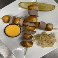 Pretzel Kabobs · Two skewers with pretzel bites and bratwurst topped with a gherkin pickle. Side of beer chee...
