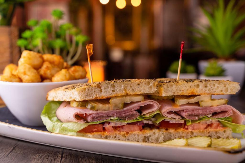 Lyon Sandwich  · Smoked Black Forest ham, salami, prosciutto, pickle, lettuce, tomato and onion, served on focaccia bread with Sriracha mayo. All sandwiches & wraps served with baked Tater Tots.