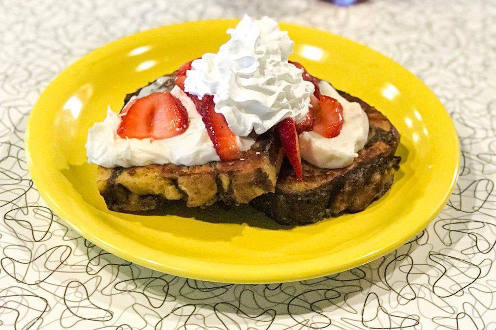 Cinnamon Roll French Toast Sweets · Two slices of grilled cinnamon french toast topped with seasonal fruit, buttercream icing, whipped cream, powdered sugar and served with maple syrup.