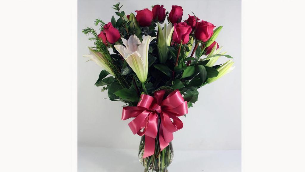 Debi Lilly Unforgettable Deluxe Arrangement · Roses, Lilies with greens in a clear garden vase with a bow. Roses can be red, pink, or yellow.