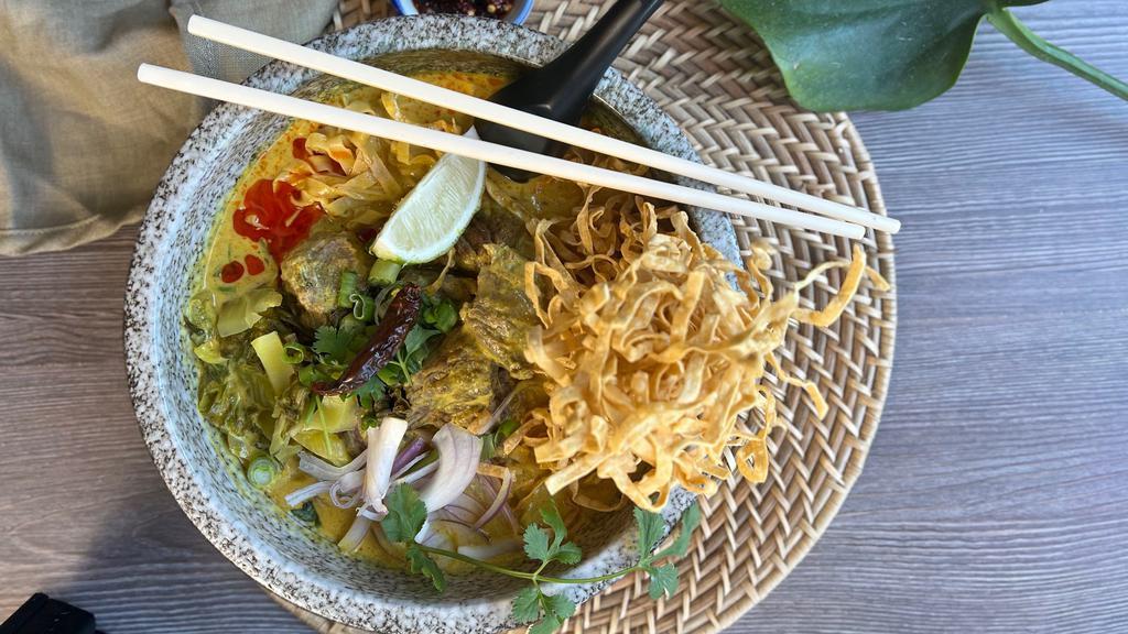 Khao Soi · Northern-style curry broth balancing between yellow curry paste and red curry paste, egg noodles topped with choice of protein, red onion, green onion, cilantro, pickled mustard greens, lime, crispy egg noodles and spicy chili oil served on the side.