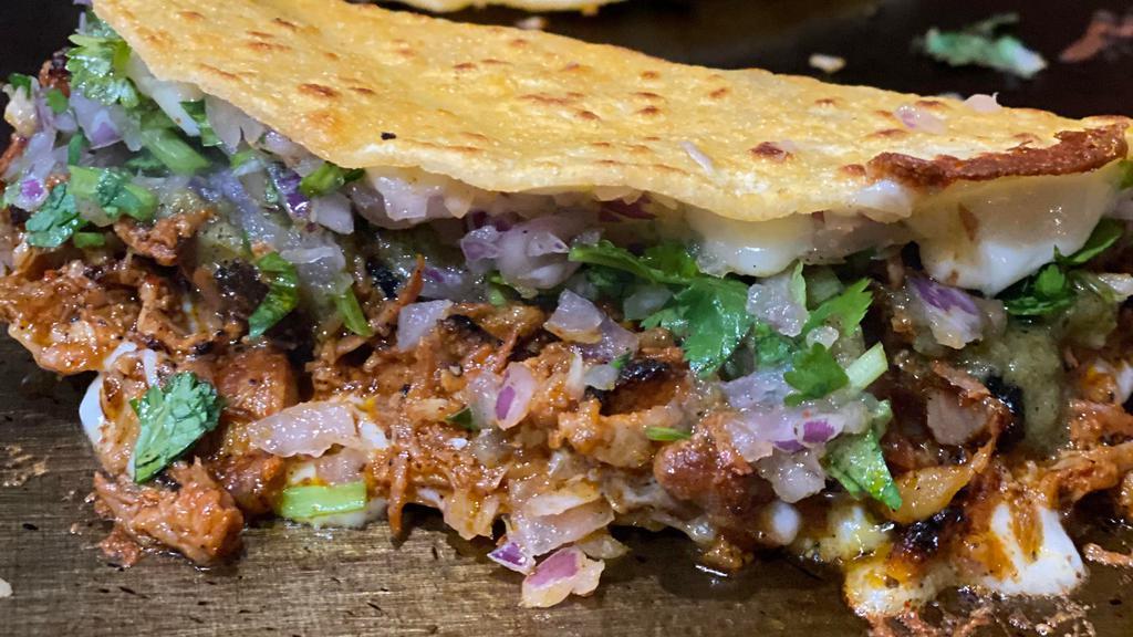 Quesa Al Pastor · Chili oil dipped corn tortilla filled with cheese, onions, cilantro, salsa verde, and Al Pastor then melted into a quesataco.  Al Pastor: Marinated and slow grilled pork and pineapple  chili oil dipped corn