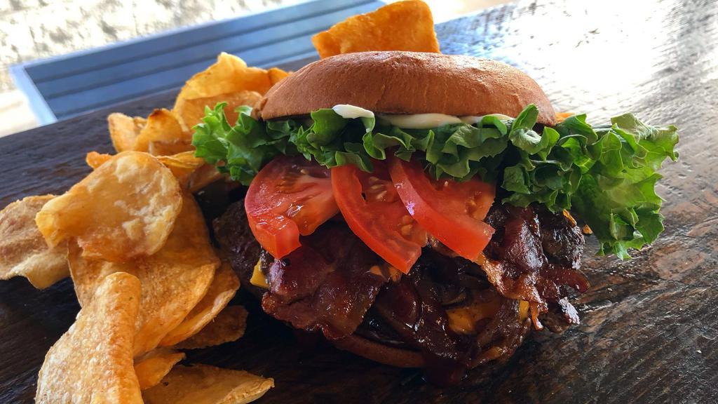 Cowboy Burger · Smashed ground beef patty with mayo, melted cheddar, tangy BBQ sauce, bacon, grilled onions, lettuce and tomato on a toasted Hawaiian bun. Served with russet potato chips.