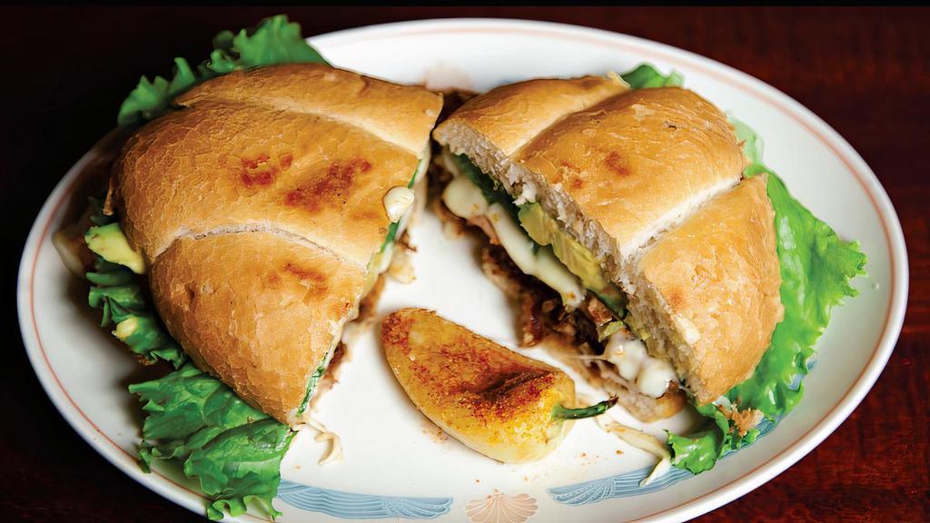 Torta Cubana · Telera bread with mayo, beans, Milanesa,  Carnitas, ham, Oaxa cheese, lettuce, tomato, avocado slices and a grill yellow pepper on  the side.