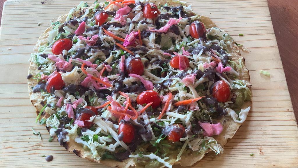 Guacamole Tlayuda · a large thin tortilla covered with guacamole spread, green cabbage, cilantro, cheese, heirloom tomatoes, pickled red onions and spice black bean salsa.