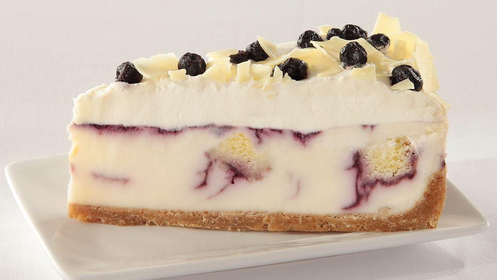Blueberry Cobbler White Chocolate Cheesecake · Creamy white chocolate cheesecake, moist chunks of vanilla bean cream cake and sweet swirls of berry compote; topped with pure whipping cream, white chocolate shavings and speckled with infused dried blueberries