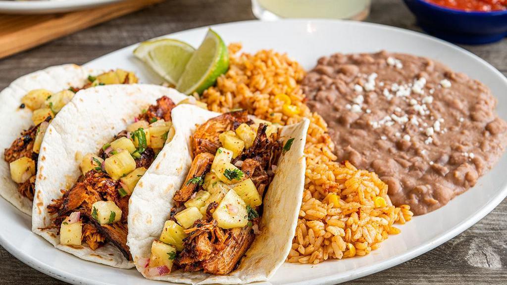 Tacos Al Pastor · Seasoned, braised carnitas and pineapple salsa in hand-pressed flour tortillas. Served with Mexican rice and choice of beans.