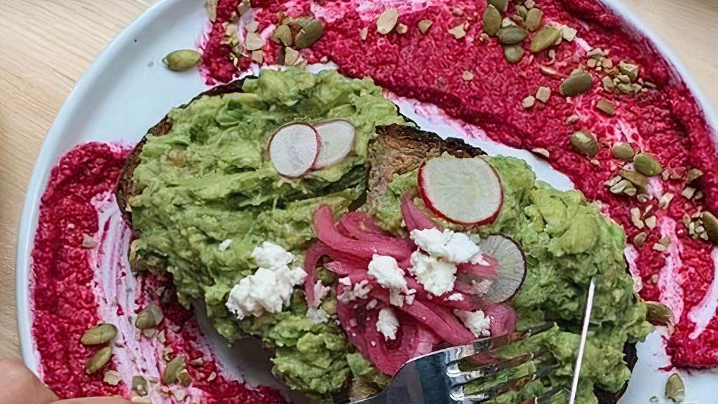 Smashing Avocado · Avocado tossed with feta, lime, cilantro on top of thick cut multigrain toast. Garnished with pickled onion, radish, pumpkin seeds and beetroot hummus