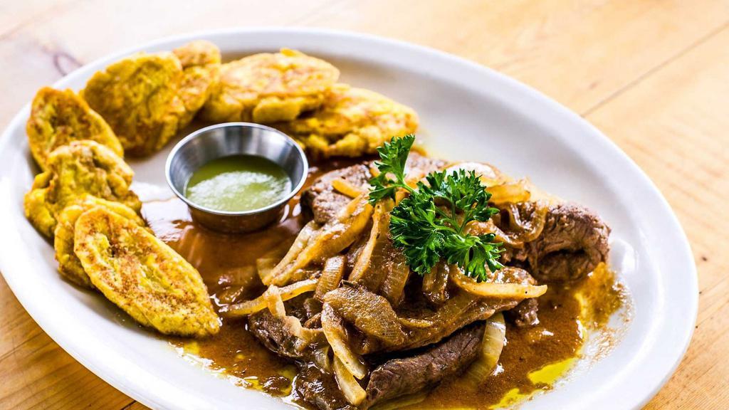 Bistec Encebollado · Marinated palomilla sirloin steak sautéed with red wine and onions, served with tostones.