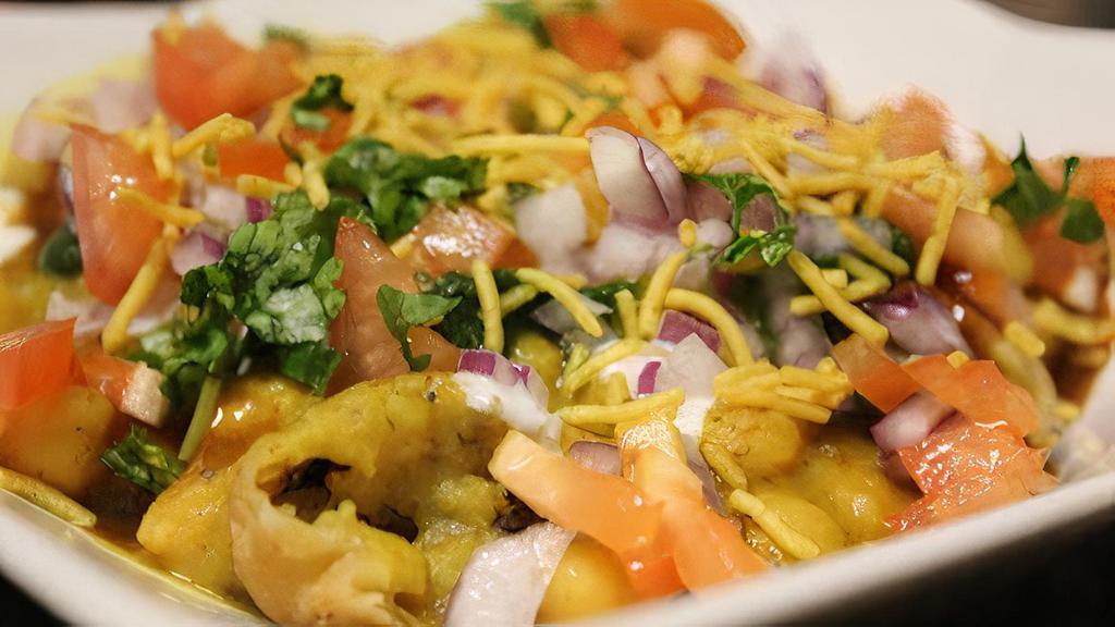 Samosa Chaat · Flattened samosas covered with chickpeas curry, chopped onions, tomatoes, coriander and chutneys.