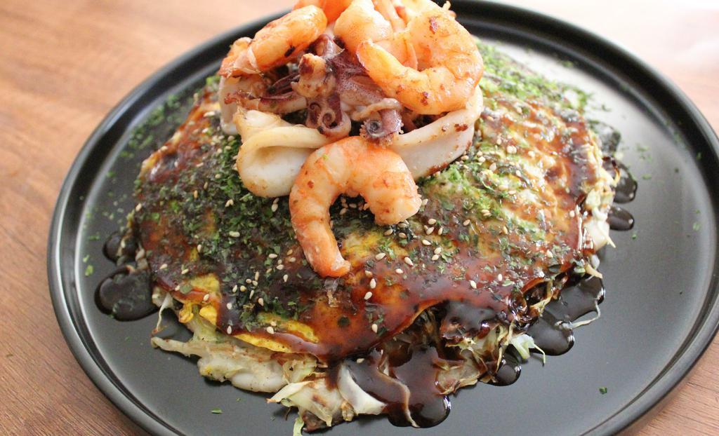 Deluxe Seafood-H · Layered  vegetables on the top of crepes  like batter with  bonito flakes, pork belly, stir fried yakisoba noodle, fried egg with Okonomiyaki sauce, aonori seaweed, sesame seeds, grilled squid and shrimp.