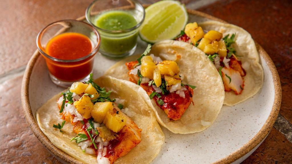 Tacos De Pescado Al Pastor (3 Pieces) · Gluten-free. Grilled fish of the day marinated in guajillo salsa, grilled pineapple, onion, and cilantro on handmade corn tortillas with salsa verde and salsa roja on the side. No mix and match.