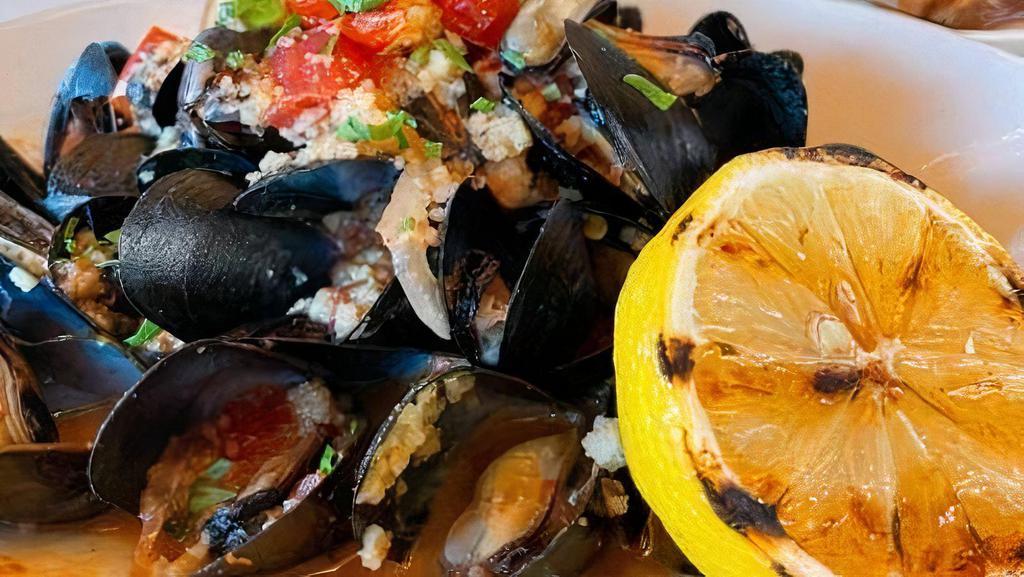 Mussels · steamed & in a spicy garlic tomato white wine sauce, topped with onion & couscous, garnished with a grilled slice of lemon.