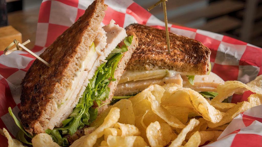 Turkey Club · Roasted turkey breast, watercress, sliced apple, lemon herb aioli, red onion jam, and whole grain bread, served with potato chips.