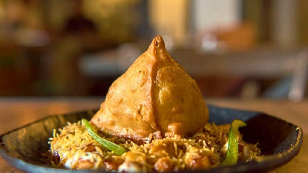 Samosa Chaat · A samosa with chickpeas, mint, tamarind, yogurt sauces, topped with chaat masala and cilantro.
