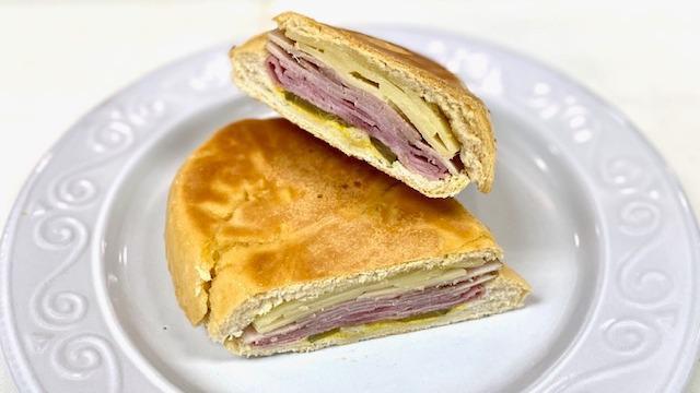 Cuban Sandwich / Sandwich Cubano (8477) · Ham, roast pork, Swiss cheese, pickles & mustard on Cuban bread roll. / Jamon ahumado, pierna de cerdo, queso suizo, pepinillos y mostaza en pan cubano.