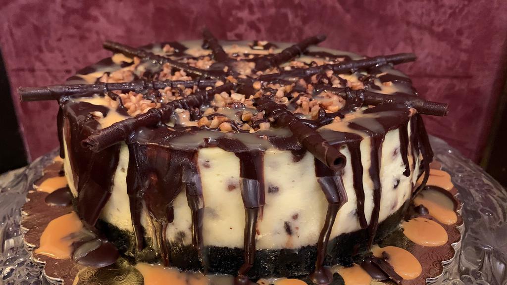 Turtle Cheesecake Slice · New York style cheesecake with an Oreo crust, baked with pecans and chocolate chips, topped with caramel and chocolate ganache.