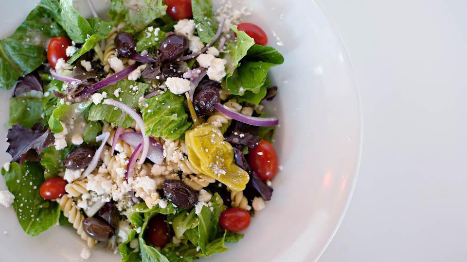 Greek Salad · Fusilli pasta, house greens, Kalamata olives, grapes tomatoes and red onions tossed in our house Greek dressing. Topped with crumbled Feta cheese and a pepperonicini pepper for an extra burst of flavor.