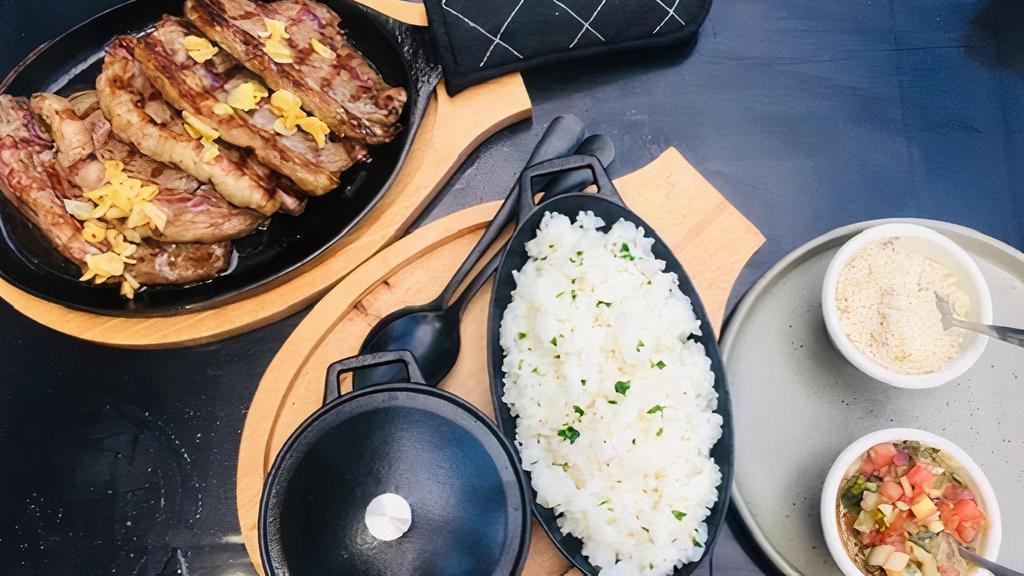 Picanha (Serves2) · char-grilled top sirloin steak cut on a sizzling skillet (5 slices), topped with roasted garlic. Served with rice, beans, farofa, and vinaigrette