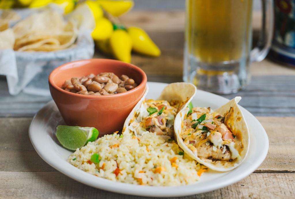 Fish Tacos · Two corn tortillas filled with blackened tilapia, shredded lettuce, and corn salsa drizzled with chipotle aioli and garnished with a lime wedge. Served with vegetable rice and charro beans.