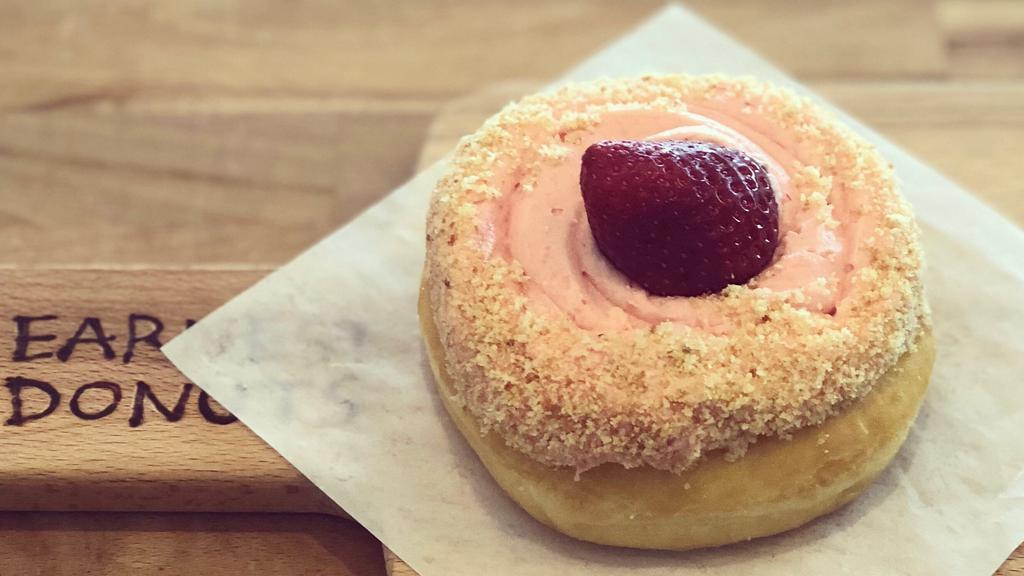 Strawberry Cheesecake · A raised donut frosted with strawberry flavored cheesecake with cake crumbs and topped with whipped cream and fresh strawberry garnish.