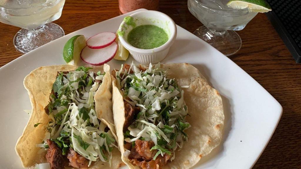 Tacos Al Pastor · Two tacos with marinated pork in a chile pineapple sauce. Topped with cilantro, onion, and salsa.