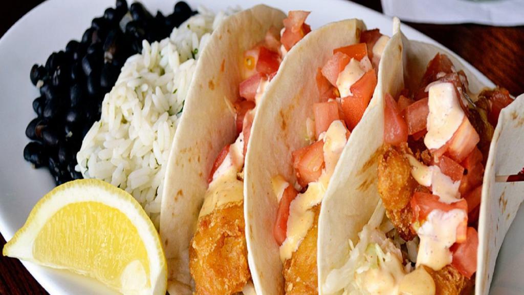 Baja Fish Tacos · Three flour tortillas filled with beer battered cod, fresh chipotle cabbage slaw, and diced tomatoes. Drizzled with lime chipotle aioli and served with black beans, cilantro lime rice, & a lemon wedge.