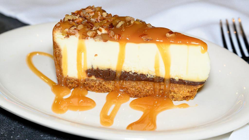 Turtle Cheesecake · New York-Style cheesecake, resting on a thick layer of fudge, covered with a rich caramel sauce, garnished with chopped.