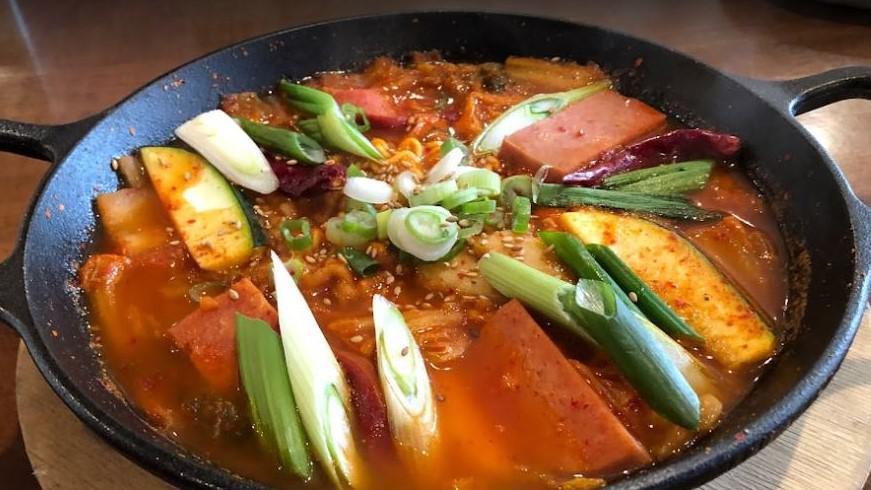 Budae Jigae / 부대찌게 · Stew loaded with kimchi, tofu, spam, sausages, ramen noodles, rice cakes, mushrooms, scallions, zucchini in spicy broth.