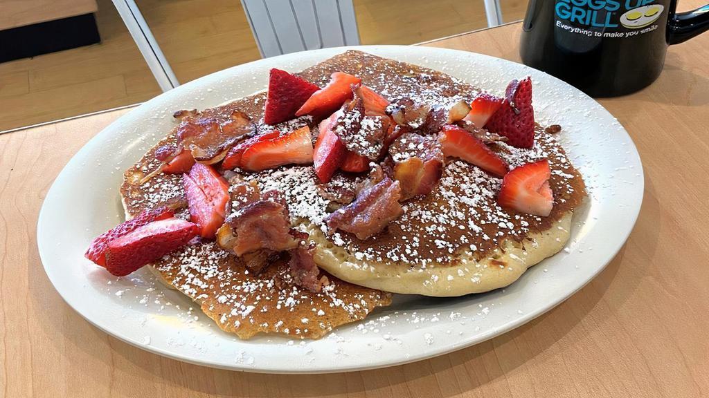 Red, White & Bacon Pancakes · Two fluffy buttermilk pancakes with crispy bacon pieces cooked into the batter, topped with fresh strawberries and powdered sugar and garnished with even more bacon.