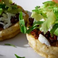 Sopes · Two corn patties topped with refried beans, shaved lettuce, green tomatillo salsa, cotija ch...
