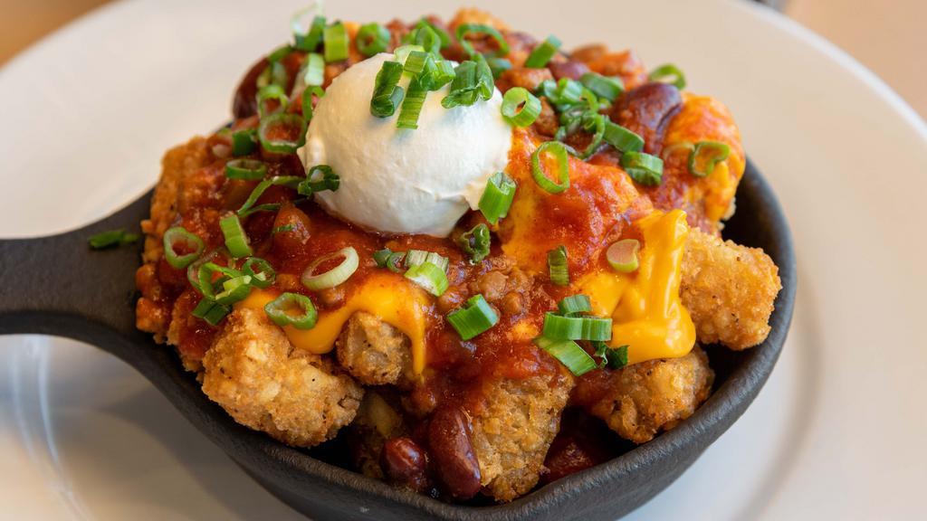 Fiesta Tot Appetizer · A skillet loaded with golden brown crispy tater tots, covered with NC’s hearty chili con carne, smooth cheddar cheese sauce, a dollop of sour cream and topped with green onions.